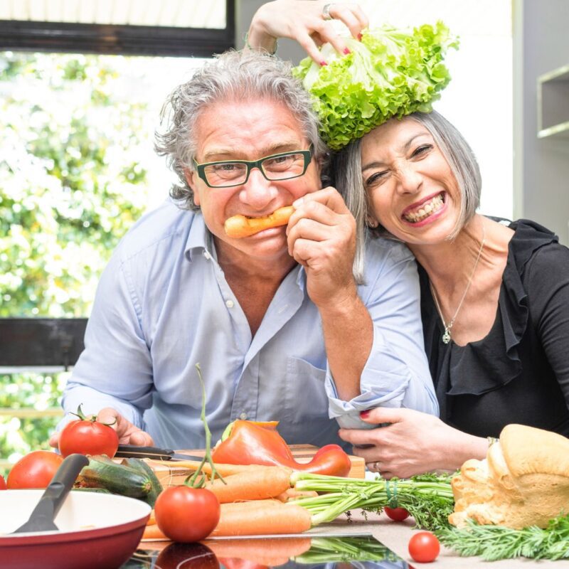 Pareja de ancianos en la cocina, sonriendo mientras preparan una comida saludable con vegetales frescos. La mujer sostiene una lechuga sobre la cabeza del hombre de forma divertida.